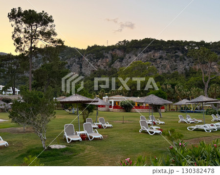 Sun loungers with straw umbrellas on a green lawn against mountain cliffs. Leisure, travel and relaxation in a natural resort environment. Sun loungers with straw umbrellas on a green lawn against mountain cliffs. Leisure, travel and relaxation in a natural resort environment. 130382478