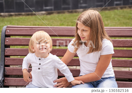 Caucasian child and teen girl sitting on a park bench smiling and enjoying sun Caucasian child and teen girl sitting on a park bench smiling and enjoying sun 130382516