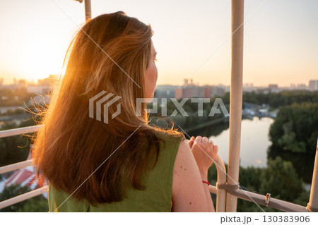 Woman with flowing hair enjoys the sunset view from a Ferris wheel, overlooking the river and city skyline 130383906