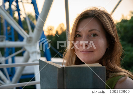 Woman holding an open book looks at the camera while sitting outdoors near a ferris wheel structure at sunset Woman holding an open book looks at the camera while sitting outdoors near a ferris wheel structure at sunset 130383909
