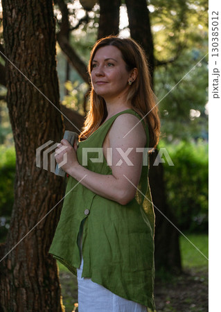 A woman in a green linen blouse and white pants stands by a tree at golden hour, holding a closed vintage book 130385012