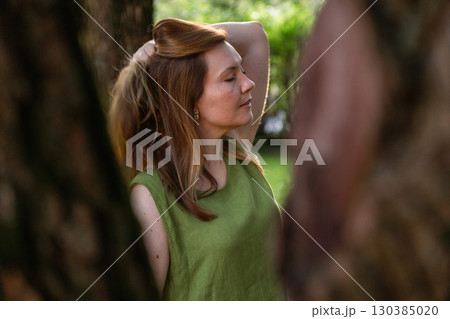 A woman in a green linen top stands among trees, her hand lifting her hair in the warm evening light 130385020