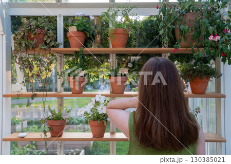 Woman with long hair in a green dress looks at clay pots with flowers on wooden shelves in a greenhouse Woman with long hair in a green dress looks at clay pots with flowers on wooden shelves in a greenhouse 130385021