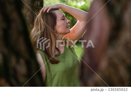 A woman in a green linen top stands among trees, her hand lifting her hair in the warm evening light A woman in a green linen top stands among trees, her hand lifting her hair in the warm evening light 130385022