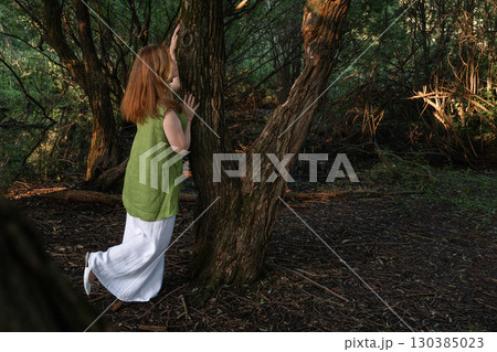 A woman in a green blouse and white trousers stands among large tree trunks, touching the bark with a calm expression 130385023