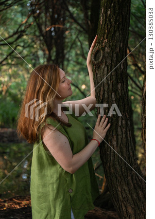 A woman in a green blouse touches a tree trunk and looks upward in a forest A woman in a green blouse touches a tree trunk and looks upward in a forest 130385038