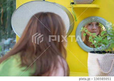 Woman in a green blouse looks at her reflection in a small round mirror framed by plants against a bright yellow wall 130385051