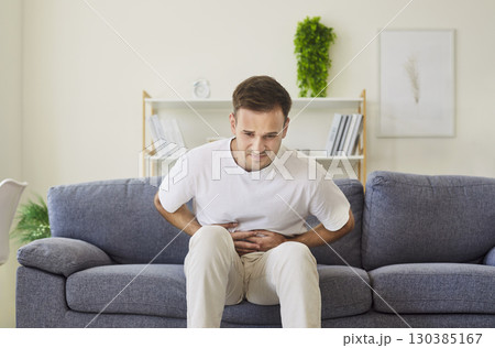 Young man hunched on couch with abdominal pain, pressing stomach in discomfort 130385167