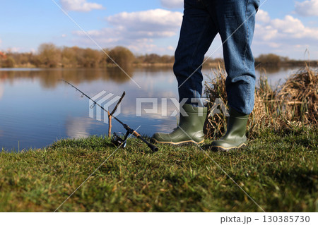 Cropped photo of fisherman's legs in rubber boots with rod, spinning reel on river bank. Unrecognisable man catching fish on lake, pond. Fishing for pike, perch on beach. Fishing day concept. 130385730