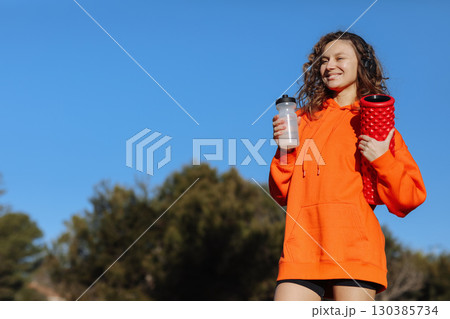 Young woman in with yoga roller drinks water from bottle, listens music in headphones, rests after workout outdoor. Fitness, sport, healthy active lifestyle. Break, hydration, wellness, aqua balance 130385734