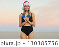 Happy slim young woman in Santa Claus hat, bikini outdoors shows smartphone, credit card, passport for book online ticket to travel on Christmas and New Year 2024 holiday vacation at tropical resort 130385756