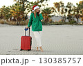 Santa woman traveler in Christmas hat with red suitcase holds passport and books online ticket by cellphone app on beach with tropical palms. Travel concept on Xmas holidays and New Year vacation 130385757