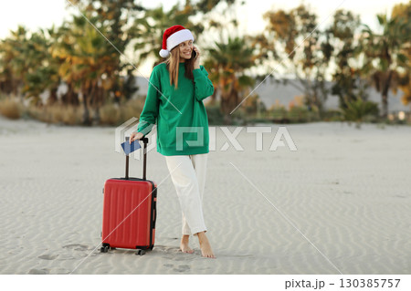 Santa woman traveler in Christmas hat with red suitcase holds passport and books online ticket by cellphone app on beach with tropical palms. Travel concept on Xmas holidays and New Year vacation 130385757