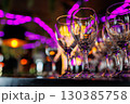 Empty clear wineglasses stand in rows on bar counter at nightclub, blurred purple neon and yellow lights on background. Clean cocktail glasses ready for celebrate party. 130385758