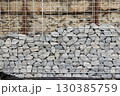 Stone fence in steel mesh and green lawn. Close up of gabion with tightly packed stones of varying sizes and textures. Combination of natural materials and industrial design. 130385759