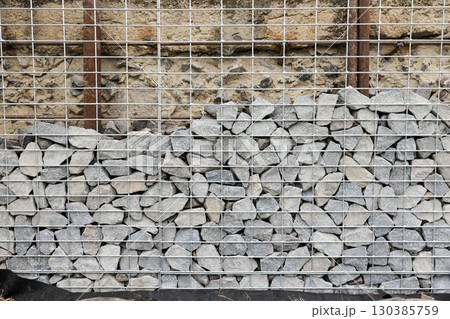 Stone fence in steel mesh and green lawn. Close up of gabion with tightly packed stones of varying sizes and textures. Combination of natural materials and industrial design. Stone fence in steel mesh and green lawn. Close up of gabion with tightly packed stones of varying sizes and textures. Combination of natural materials and industrial design. 130385759