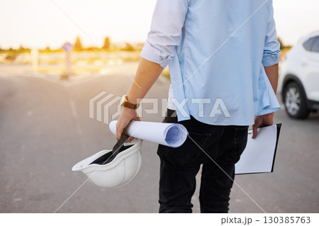 Unrecognisable engineer, architect or worker holds in hand protective white hard hat and blueprint on multi-apartment residential complex background, sunlighting. Concept of new real estate. Unrecognisable engineer, architect or worker holds in hand protective white hard hat and blueprint on multi-apartment residential complex background, sunlighting. Concept of new real estate. 130385763