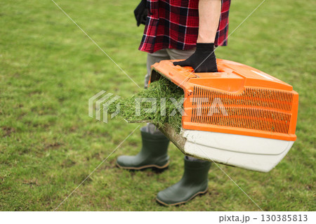 Unrecognisable man in casual clothes mows lawn with lawn mower at own backyard, holds bag with cut grass. Husband takes care of garden. Modern gasoline garden equipment. Landscaping work at home Unrecognisable man in casual clothes mows lawn with lawn mower at own backyard, holds bag with cut grass. Husband takes care of garden. Modern gasoline garden equipment. Landscaping work at home 130385813