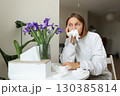 Unhappy sick young woman with paper tissues inside nose from runny nose and sneezes sits by table at home. Female with pollen allergy, flu, cold, rhinitis, sinusitis and with viral infection 130385814