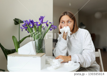 Unhappy sick young woman with paper tissues inside nose from runny nose and sneezes sits by table at home. Female with pollen allergy, flu, cold, rhinitis, sinusitis and with viral infection 130385814