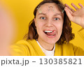 Playful young woman takes a selfie, smiles, sticks out her tongue, holds glasses on yellow backdrop. Modern lifestyle, social media, youth influencer marketing, self expression, personal branding 130385821
