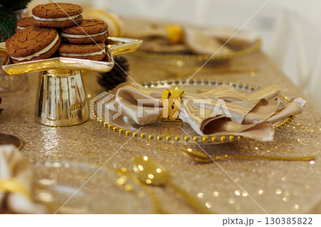 Luxury golden Christmas dinner table setting. Glowing tablecloth, napkin with steel napkin ring on glass plate, cutlery, candlesticks. Xmas tree illuminating on backdrop. Festive new 2026 year party. 130385822