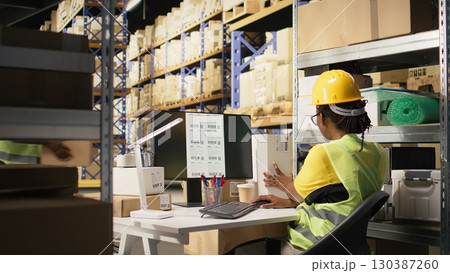 Storehouse black worker reviewing digital shipment status, using awb numbers on tracking software. Woman handling data driven order tracking and delivery updates in warehouse. Camera A. Storehouse black worker reviewing digital shipment status, using awb numbers on tracking software. Woman handling data driven order tracking and delivery updates in warehouse. Camera A. 130387260