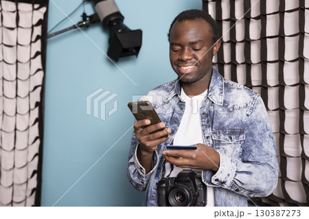 African American man standing in his studio, happily making an online purchase with his cellphone. Cheerful male photographer entering his credit card details on his mobile, completing a transaction. African American man standing in his studio, happily making an online purchase with his cellphone. Cheerful male photographer entering his credit card details on his mobile, completing a transaction. 130387273