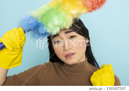 Exhausted asian female janitor touching head with feather duster, feeling overwhelmed by cleaning duties. Tired housekeeper with yellow gloves, showing signs of fatigue after long day of chores. 130387297