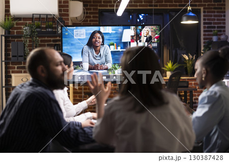 Multiethnic colleagues discussing startup project plans in late night meeting. A video conference with remote team is visible on digital screen, symbolizing modern business collaboration. 130387428