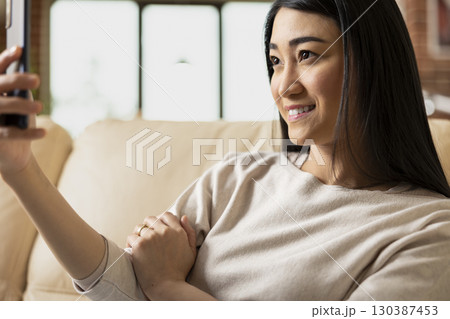 Smiling female entrepreneur sits comfortably on couch, snapping selfie with cellphone. Joyful remote worker enjoying personal time, documenting day for social media in brick wall apartment. 130387453