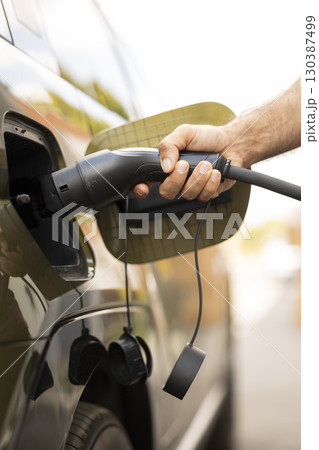 Close up of man connecting cable to vehicle inlet port of zero emissions EV car to charge it while parked. Driver using green tech electric vehicle socket to charge battery 130387499