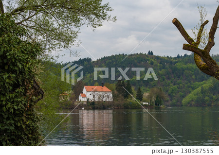 White church with red roof by forested riverbank - peaceful spring landscape with cloudy sky 130387555