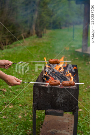 Grilling sausages on skewers over open flame in metal barbecue grill during outdoor cookout . 130387556