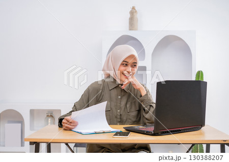 A Young Woman Engaged in Work at Home Utilizing a Laptop Alongside Documents on Her Desk 130388207