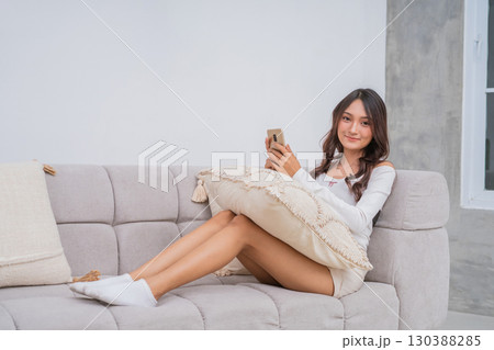 A young woman relaxes on a couch with her phone, surrounded by decorative pillows A young woman relaxes on a couch with her phone, surrounded by decorative pillows 130388285