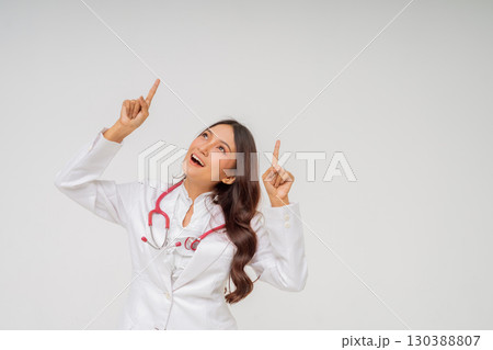 A cheerful doctor in a white coat points upward with a stethoscope, showing expertise A cheerful doctor in a white coat points upward with a stethoscope, showing expertise 130388807