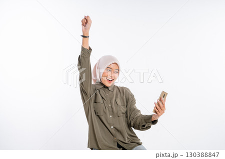 A Cheerful Young Woman is Joyfully Celebrating while Holding a Smartphone in Her Hand 130388847