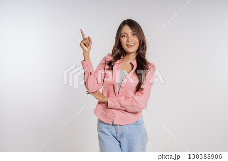 A cheerful young woman in a casual outfit is smiling and pointing upwards 130388906