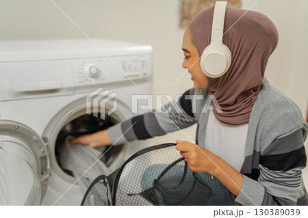 A Young Woman Happily Doing Laundry with Headphones in a Cozy Home Setting 130389039