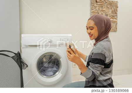 A young woman is happily using her smartphone while doing laundry at home today A young woman is happily using her smartphone while doing laundry at home today 130389066