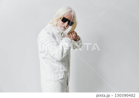 Stylish elderly man with white hair and sunglasses wearing a white outfit, confidently posing in a modern studio with a minimalistic gray background 130390482