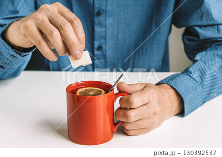 person adds sugar cube to hot tea with lemon slice in red mug during cozy indoor moment. closeup. 130392537