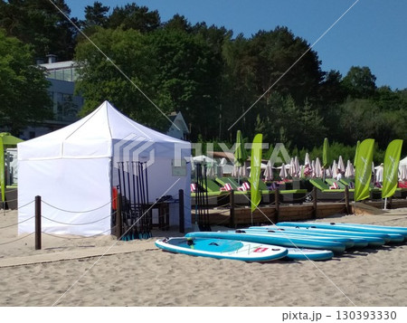 Sandy beach area with a row of blue stand-up paddleboards lined up near a white canopy tent. Paddles are propped against a wooden post. Yurmala beach, Latvia Sandy beach area with a row of blue stand-up paddleboards lined up near a white canopy tent. Paddles are propped against a wooden post. Yurmala beach, Latvia 130393330