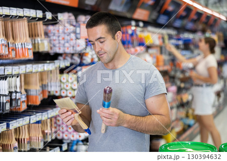 Focused young man choosing brushes in painting supplies store 130394632