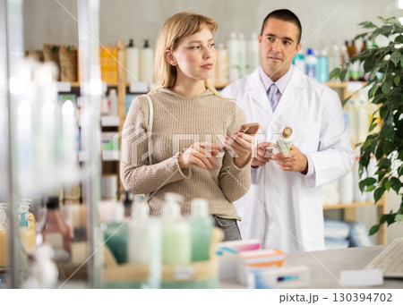 Girl client scrolls phone and shopping in pharmacy, male pharmacist in background. 130394702