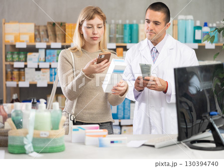 Girl in pharmacy scans QR code on emollient for sensitive skin, male pharmacist in background 130394740