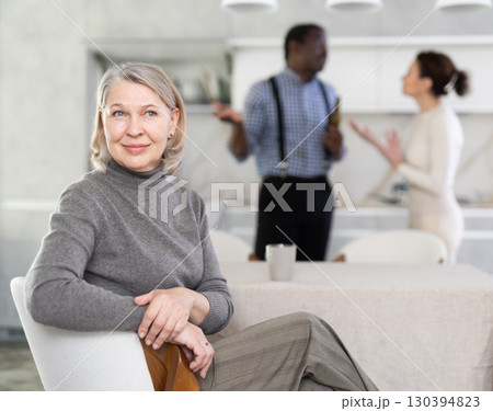 Smiling old woman sitting at the kitchen table with her back to couple quarreling together Smiling old woman sitting at the kitchen table with her back to couple quarreling together 130394823