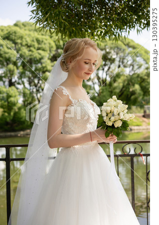 Beautiful bride stands by serene lake holding a bouquet of white flowers 130395275