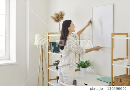 Cheerful woman arranging piece of art on wall in her sunny apartment Cheerful woman arranging piece of art on wall in her sunny apartment 130396692